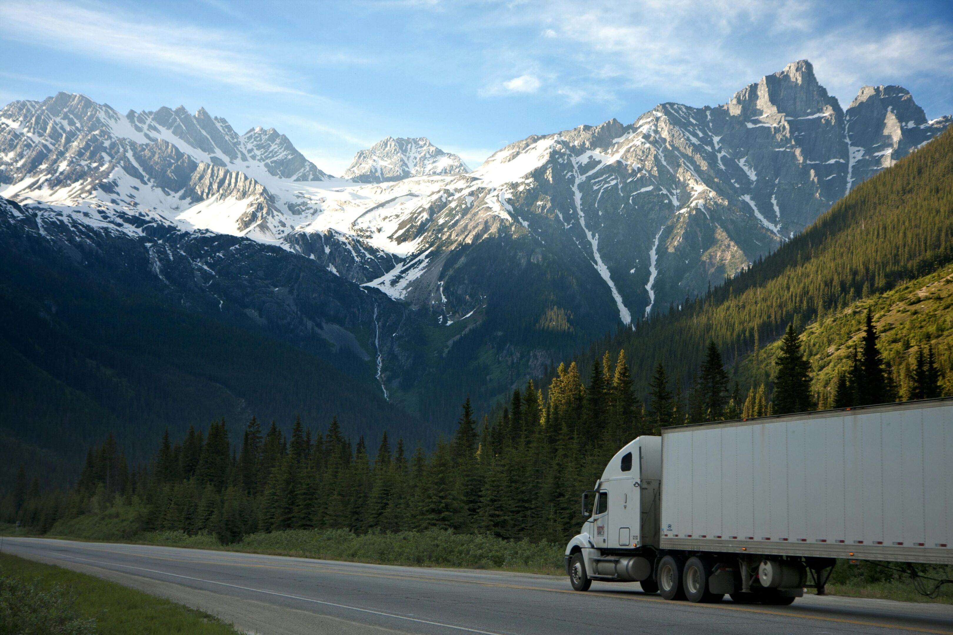 A white semi truck drives on a mountain road beneath snowy peaks.