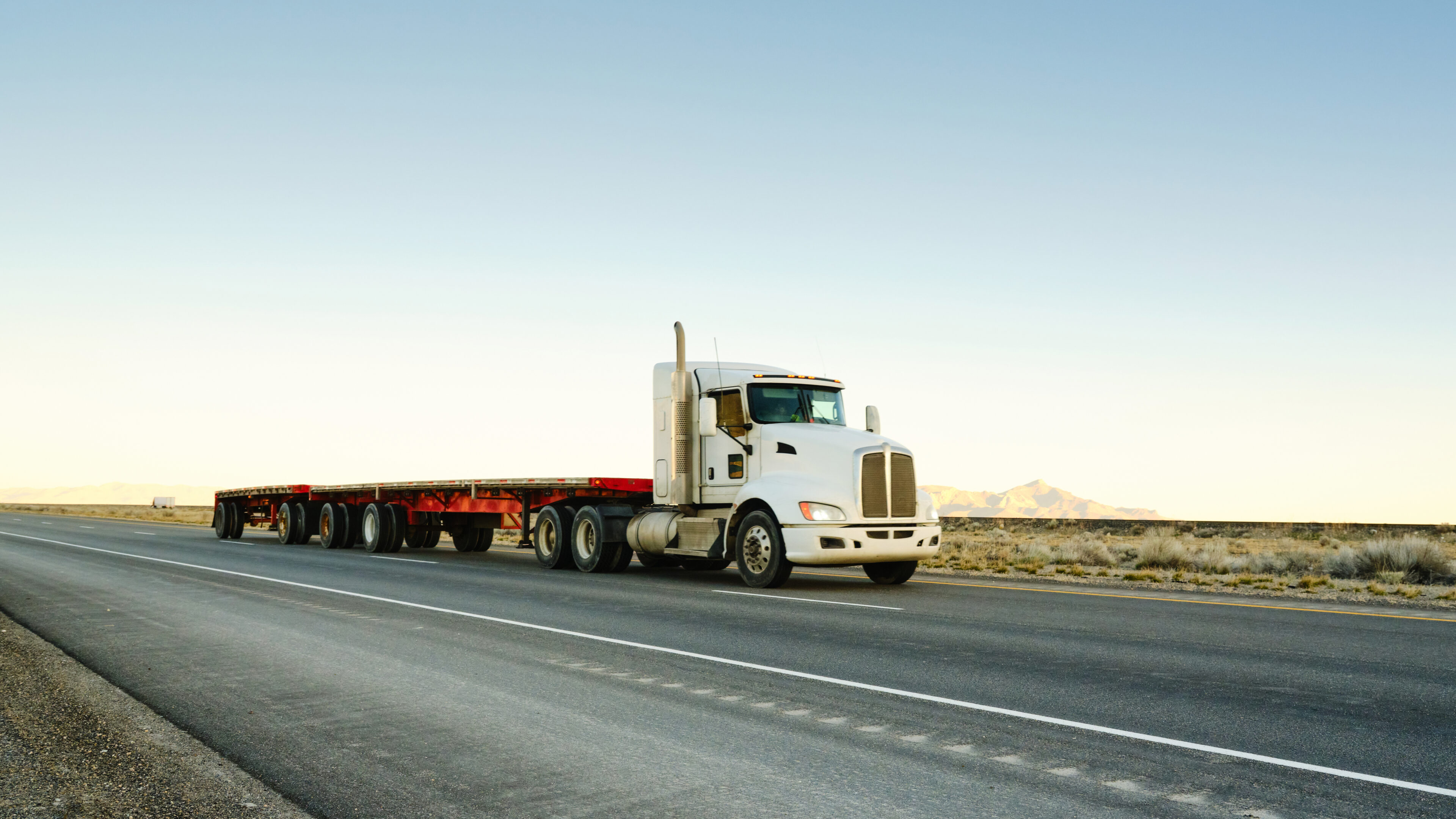 White semi-truck with flatbed trailer driving on an open highway.