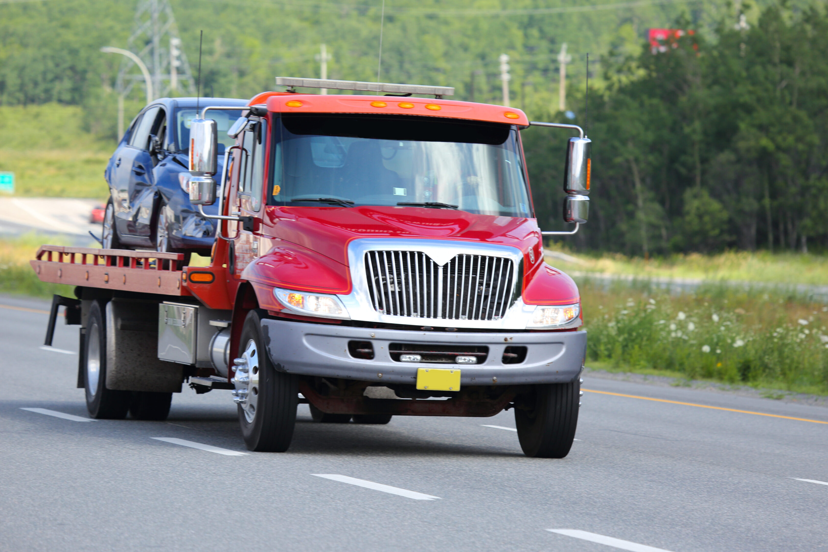 Red tow truck transporting a damaged blue car on the highway.
